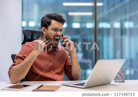 Angry businessman shouting on phone at office desk expressing frustration. Young professional arguing call with clenched fist, laptop, and tablet on desk. Office environment with blurred background. 117140713