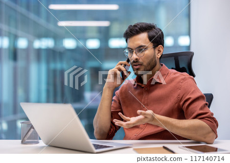 Businessman talking on phone while working on laptop in modern office setting. Professional engaged in conversation, multitasking in workspace. Concept of communication, technology, and business. Businessman talking on phone while working on laptop in modern office setting. Professional engaged in conversation, multitasking in workspace. Concept of communication, technology, and business. 117140714