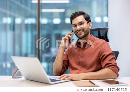 Smiling businessman in glasses talking on phone while working at desk with laptop in modern office. Concept of business communication and professional workspace Smiling businessman in glasses talking on phone while working at desk with laptop in modern office. Concept of business communication and professional workspace 117140715