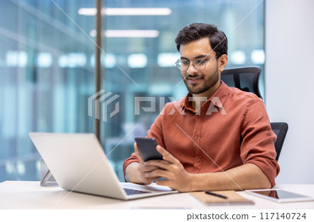 Young professional with glasses working at desk in modern office using smartphone and laptop, displaying productivity, technology, and communication. Image portrays a comfortable work environment with Young professional with glasses working at desk in modern office using smartphone and laptop, displaying productivity, technology, and communication. Image portrays a comfortable work environment with 117140724