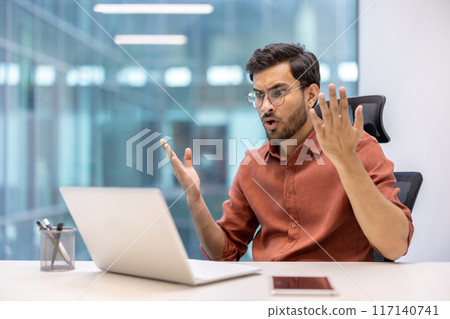 Office worker expressing frustration in modern workspace, sitting at desk with laptop and smartphone. Man showing gestures of disbelief and confusion in professional environment. 117140741