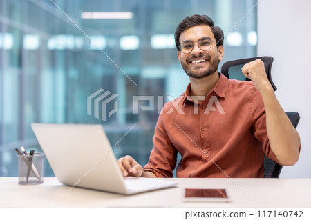 Smiling young professional celebrating success in modern office with laptop and phone on desk. Man expressing joy and achievement in business environment. Smiling young professional celebrating success in modern office with laptop and phone on desk. Man expressing joy and achievement in business environment. 117140742