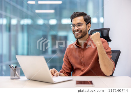 Smiling office worker in casual attire celebrating success at desk with laptop. Professional sitting in modern office environment, expressing happiness and achievement. 117140744