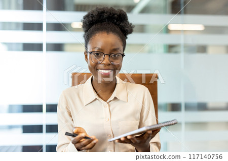 Confident businesswoman smiling during office video call. Professional holding notepad pen, ready for meeting. Modern office setting enhances corporate ambiance. Concept of remote work communication Confident businesswoman smiling during office video call. Professional holding notepad pen, ready for meeting. Modern office setting enhances corporate ambiance. Concept of remote work communication 117140756