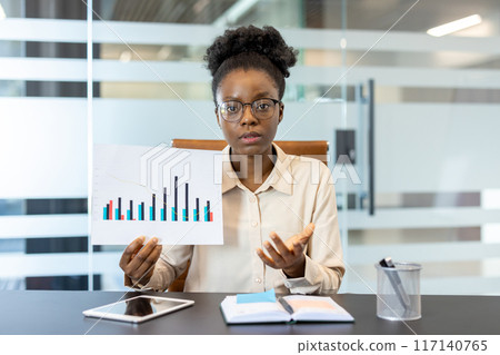 Businesswoman holding graph chart discussing financial performance during video call. Professional using remote communication for business presentation. Modern office setting with technology tools. 117140765