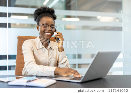 Young African American businesswoman working in office. Employee on phone and laptop smiling, wearing glasses and white shirt at desk. Professional workplace environment Young African American businesswoman working in office. Employee on phone and laptop smiling, wearing glasses and white shirt at desk. Professional workplace environment 117141030