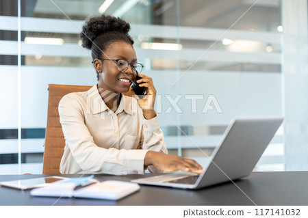 Smiling businesswoman in glasses talking on phone while working on laptop in modern office. Image portrays professionalism, productivity, and modern workplace environment. 117141032