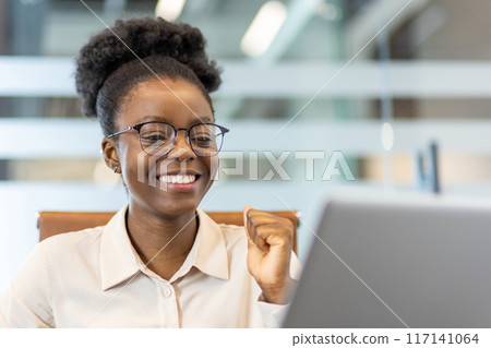 Happy businesswoman with glasses celebrating success while working on computer in modern office. Confident and successful female professional enjoying achievement or milestone at work. 117141064