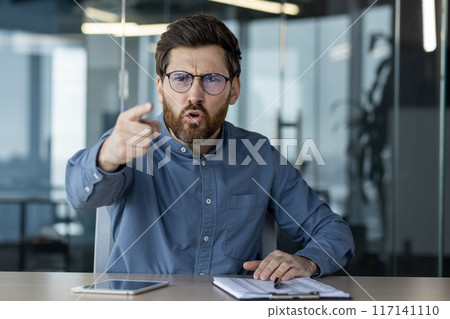 Angry businessman in glasses pointing finger during office meeting. Man expressing frustration or disagreement while discussing issues. Digital tablet and paperwork on desk. 117141110