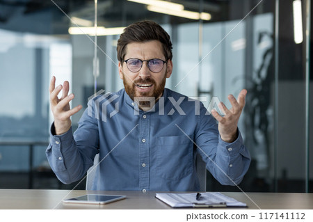 Portrait of an angry young man sitting at an office desk and expressively talking to the camera, gesturing with his hands. Portrait of an angry young man sitting at an office desk and expressively talking to the camera, gesturing with his hands. 117141112