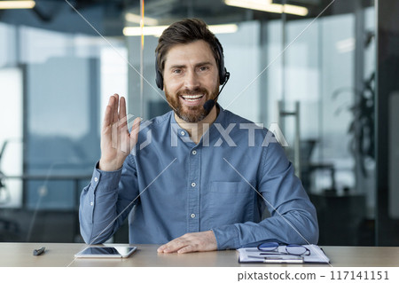 A smiling young man in a headset sits in the office, talks, smiles and greets with his hand to the camera, conducts online consultations and remote training. A smiling young man in a headset sits in the office, talks, smiles and greets with his hand to the camera, conducts online consultations and remote training. 117141151