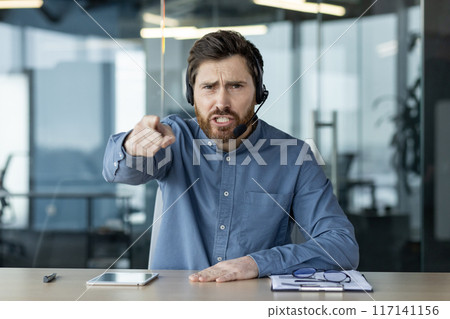 Portrait of a young man in a headset sitting in the office at the table, talking emotionally online, shouting and pointing at the camera. Portrait of a young man in a headset sitting in the office at the table, talking emotionally online, shouting and pointing at the camera. 117141156