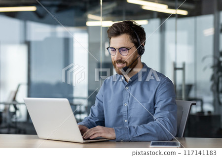 A smiling young man in a blue shirt is sitting at a desk in a modern office, wearing a headset, working and studying at a laptop. 117141183