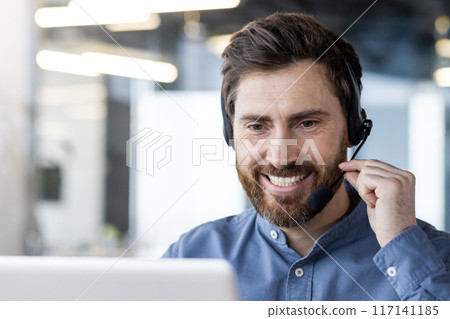 Close-up photo of a young smiling man wearing a headset sitting in the office in front of a laptop ,and talking on a video call. 117141185
