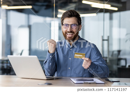 Portrait of a happy young man businessman who is happy about financial success, sitting in the office at the table, holding a credit card in his hands and showing a victory gesture with his hand. Portrait of a happy young man businessman who is happy about financial success, sitting in the office at the table, holding a credit card in his hands and showing a victory gesture with his hand. 117141197