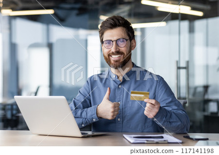 Portrait of a young smiling man sitting in the office at the table, holding and showing a golden credit card in his hand, showing the camera a super success sign with his finger. 117141198