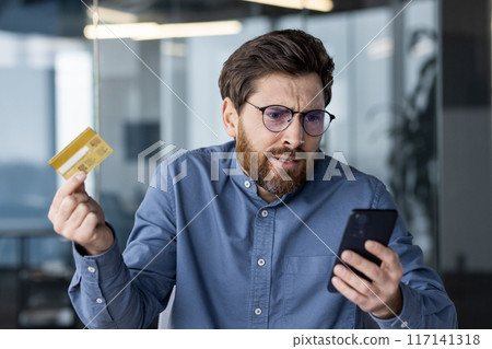 Close-up photo of a shocked young man in glasses and a blue shirt who is in the office, holding a credit card and looking disappointed at the mobile phone screen. Close-up photo of a shocked young man in glasses and a blue shirt who is in the office, holding a credit card and looking disappointed at the mobile phone screen. 117141318