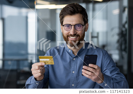 Close-up portrait of young man in glasses and shirt sitting in modern office at workplace, holding credit card and mobile phone, smiling and looking at camera. Close-up portrait of young man in glasses and shirt sitting in modern office at workplace, holding credit card and mobile phone, smiling and looking at camera. 117141323