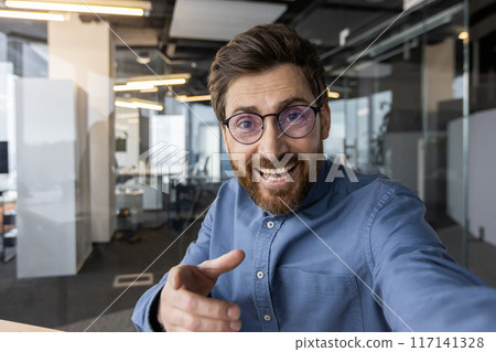 Close-up portrait of a young smiling man in glasses and a shirt standing in a modern office and chatting online while pointing, his finger at the camera. Close-up portrait of a young smiling man in glasses and a shirt standing in a modern office and chatting online while pointing, his finger at the camera. 117141328