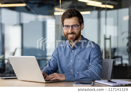 Portrait of a young man in glasses sitting at a desk in the office and working on a laptop, looking and smiling at the camera. 117141329