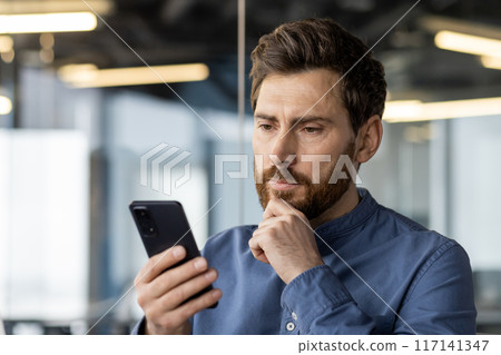 Close-up photo of a thoughtful serious young man who is in the office and reads a message, looks at the mobile phone screen, holds his hand by his beard. 117141347