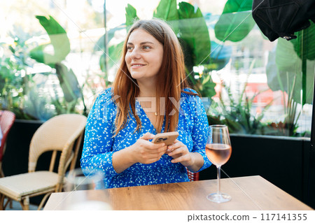 White wine glass in an outdoor cafe. Happy Woman holding glass with wine and using mobile phone outdoors White wine glass in an outdoor cafe. Happy Woman holding glass with wine and using mobile phone outdoors 117141355