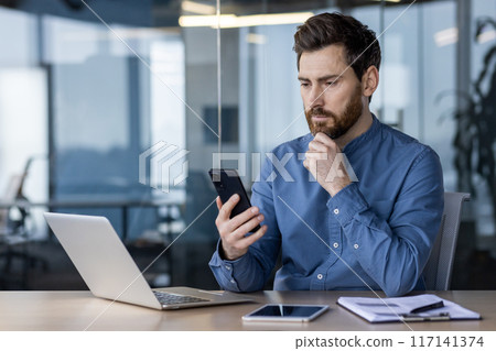 A serious and pensive young man is sitting in the office at a desk with a laptop and looking focused at the mobile phone he is holding in his hands. 117141374