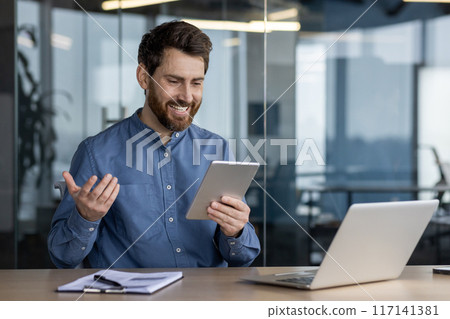 A smiling young man works in the office, sits at a desk with a laptop, holds a tablet in his hand and talks on a video call. A smiling young man works in the office, sits at a desk with a laptop, holds a tablet in his hand and talks on a video call. 117141381