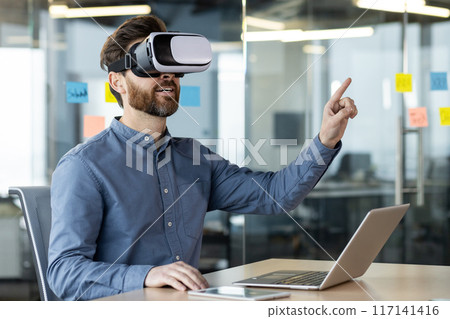 Businessman using virtual reality headset working on laptop in office. Exploring VR technology, innovative interaction, workplace creativity. Casual wear reflecting modern work environment 117141416