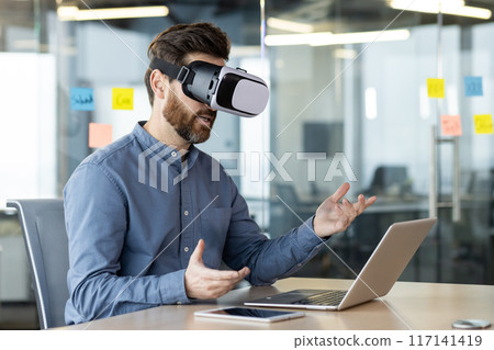 A young businessman works in the office, sits at a table with a laptop, wears a virtual mask and communicates online via video communication, gesturing with his hands. 117141419
