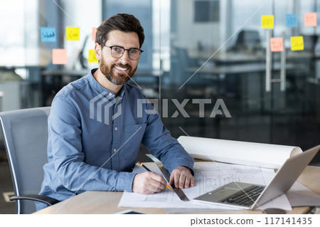 Smiling architect in blue shirt working on blueprints at office desk with laptop and sticky notes in background. Concept of planning, design, construction, and modern workplace environment. Smiling architect in blue shirt working on blueprints at office desk with laptop and sticky notes in background. Concept of planning, design, construction, and modern workplace environment. 117141435