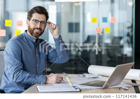 Portrait of a young smiling male developer, architect, who works in the office with a laptop and with planning documents, looks at the camera and holds glasses with his hand. Portrait of a young smiling male developer, architect, who works in the office with a laptop and with planning documents, looks at the camera and holds glasses with his hand. 117141436