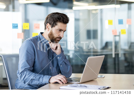 A serious and focused young man is sitting at a desk in the office and working on a laptop, looking thoughtfully at the screen. A serious and focused young man is sitting at a desk in the office and working on a laptop, looking thoughtfully at the screen. 117141438