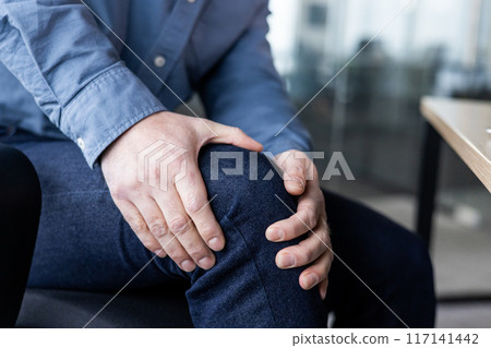 Close-up of person holding knee indicating discomfort from pain or injury. Office environment in background suggesting potential workplace-related health issues or stress. Focus on joint pain 117141442