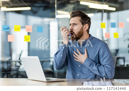 A young man businessman atsmatic sits in the office at the table, holds his hand to his chest and uses an aerosol spray. A young man businessman atsmatic sits in the office at the table, holds his hand to his chest and uses an aerosol spray. 117141448