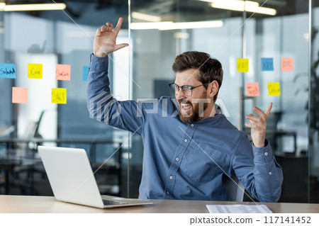 Man celebrating success in office while working on laptop surrounded by sticky notes. Happiness and accomplishment during work. Modern office vibe. Man celebrating success in office while working on laptop surrounded by sticky notes. Happiness and accomplishment during work. Modern office vibe. 117141452