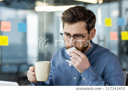 Close-up photo of a sick young male office worker sitting at his workplace, holding a cup of hot drink and medicine in his hand, wiping his nose from a runny nose with a napkin. Close-up photo of a sick young male office worker sitting at his workplace, holding a cup of hot drink and medicine in his hand, wiping his nose from a runny nose with a napkin. 117141453