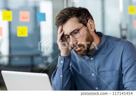 Tired and worried young businessman man sitting at workplace in front of laptop and upset with hand on head. Close-up photo. 117141454