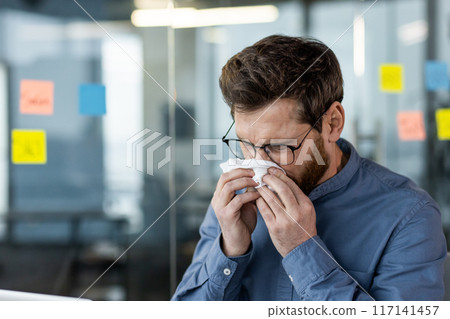 Businessman sneezing into tissue in modern office environment. Man wearing glasses suffering from cold or allergies. Concept of illness, workplace health, and hygiene. 117141457