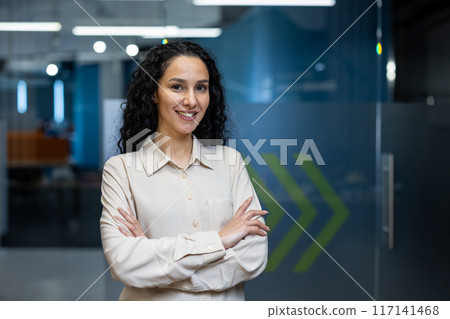 Confident businesswoman with curly hair standing with arms crossed in modern office. Professional appearance and positive expression. Concept of leadership, success, and workplace environment. Confident businesswoman with curly hair standing with arms crossed in modern office. Professional appearance and positive expression. Concept of leadership, success, and workplace environment. 117141468
