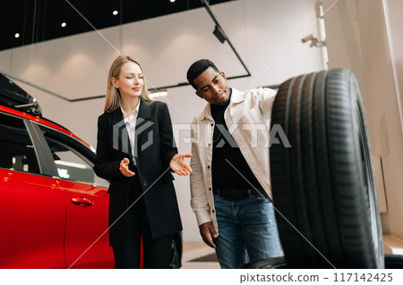 Low-angle view of saleswoman in business suit talking about characteristic of tires product to African-American customer young man came to look at assortment represented in auto service shop. 117142425