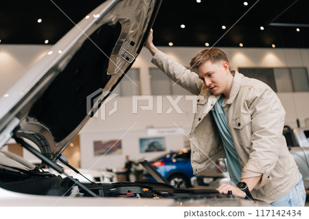Buyer male checking motor of new luxury car under hood in auto dealership before purchasing. Young man customer walking in modern auto showroom and choosing new vehicle to buy or rent. 117142434