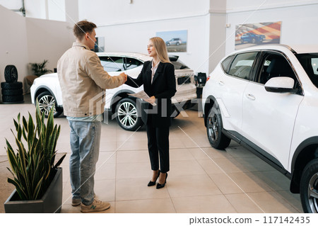 Full length portrait of female manager in suit giving car keys to client, congratulating male buyer with purchase in auto showroom. Saleswoman and male customer shaking hands after successful deal Full length portrait of female manager in suit giving car keys to client, congratulating male buyer with purchase in auto showroom. Saleswoman and male customer shaking hands after successful deal 117142435