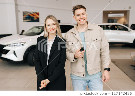 Happy young man buyer holding in hand keys of SUV car standing posing with friendly blond saleswoman in suit in bright showroom, smiling looking at camera. Concept of buying new modern vehicle. 117142436