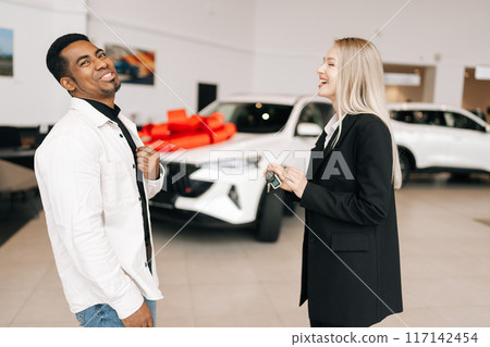 Friendly car saleswoman in business suit telling cheerful black male buyer about luxurious car in motor show. Laughing African-American client looking at auto and listening to dealer. 117142454
