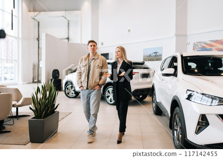 Wide shot of stylish female manager in suit showing new cars to male buyer and walking in modern showroom spbas. Young woman car dealer talking to man client and showing models of transport. Wide shot of stylish female manager in suit showing new cars to male buyer and walking in modern showroom spbas. Young woman car dealer talking to man client and showing models of transport. 117142455