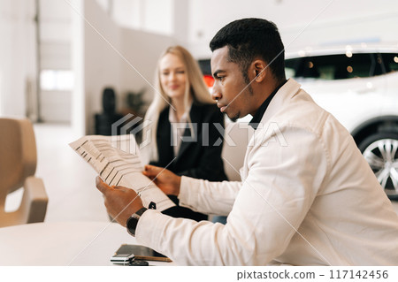Side view of focused African-American male customer carefully reading car purchase agreement before signing in dealership office. Positive female car dealer consulting doubtful buyer at showroom. Side view of focused African-American male customer carefully reading car purchase agreement before signing in dealership office. Positive female car dealer consulting doubtful buyer at showroom. 117142456