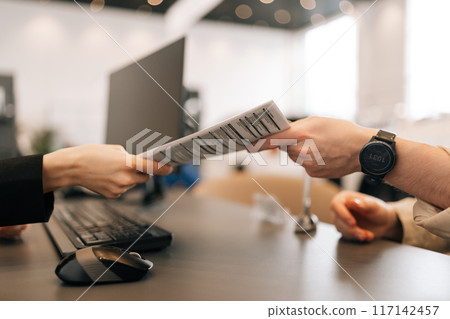 Close-up hands of unrecognizable manager and client exchanging document sitting at desk in office. Female manager handing over contract to be read and signed at business meeting, closeup side view. Close-up hands of unrecognizable manager and client exchanging document sitting at desk in office. Female manager handing over contract to be read and signed at business meeting, closeup side view. 117142457