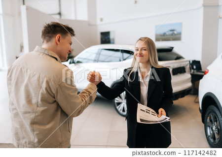 Portrait of funny saleswoman in business suit and happy customer male shaking hand in auto dealership. Happy smiling car owner during handing over keys at auto showroom. Concept of buying new auto. 117142482