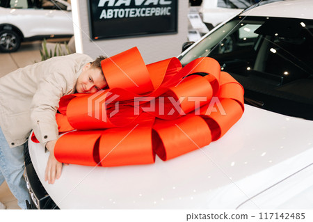 Joyful male customer lying on white luxury new car with large red bow on hood and smiling with closed eyes. Expensive purchase of cheerful male client person. Concept of buying new auto at showroom. Joyful male customer lying on white luxury new car with large red bow on hood and smiling with closed eyes. Expensive purchase of cheerful male client person. Concept of buying new auto at showroom. 117142485